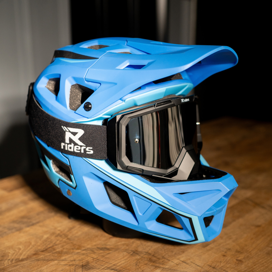 A bright blue full-face mountain bike helmet with a matching visor and goggles, resting on a wooden surface. The helmet features black vents, a black chin strap with a white 'Riders' logo, and a black visor frame.